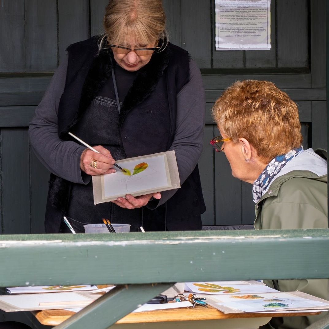 A woman demonstrating painting techniques on a botanical illustration to two seated individuals at a table inside a wooden shelter.