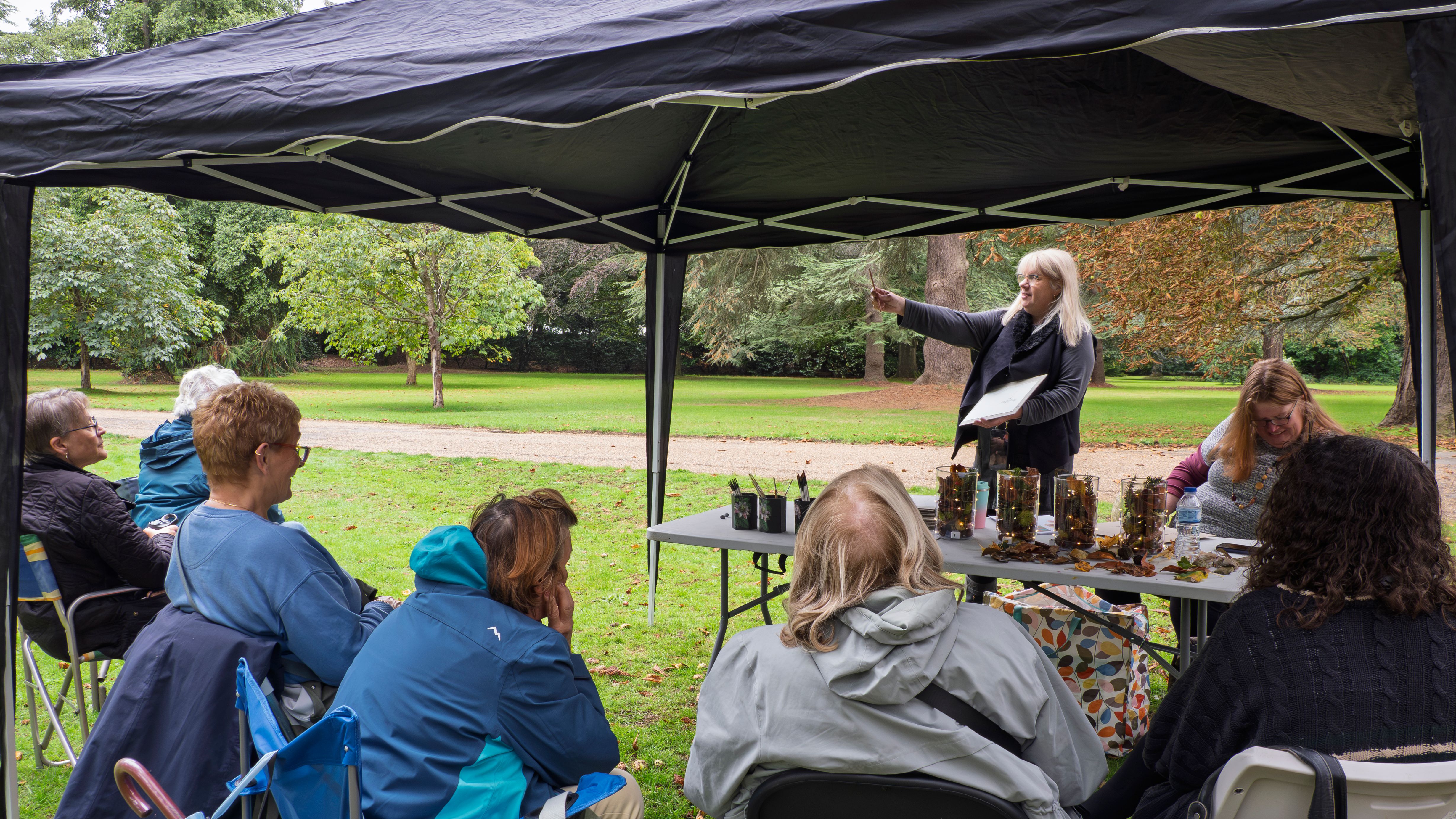 A group of people sitting under a canopy tent on a lawn, listening to a woman give a presentation.
