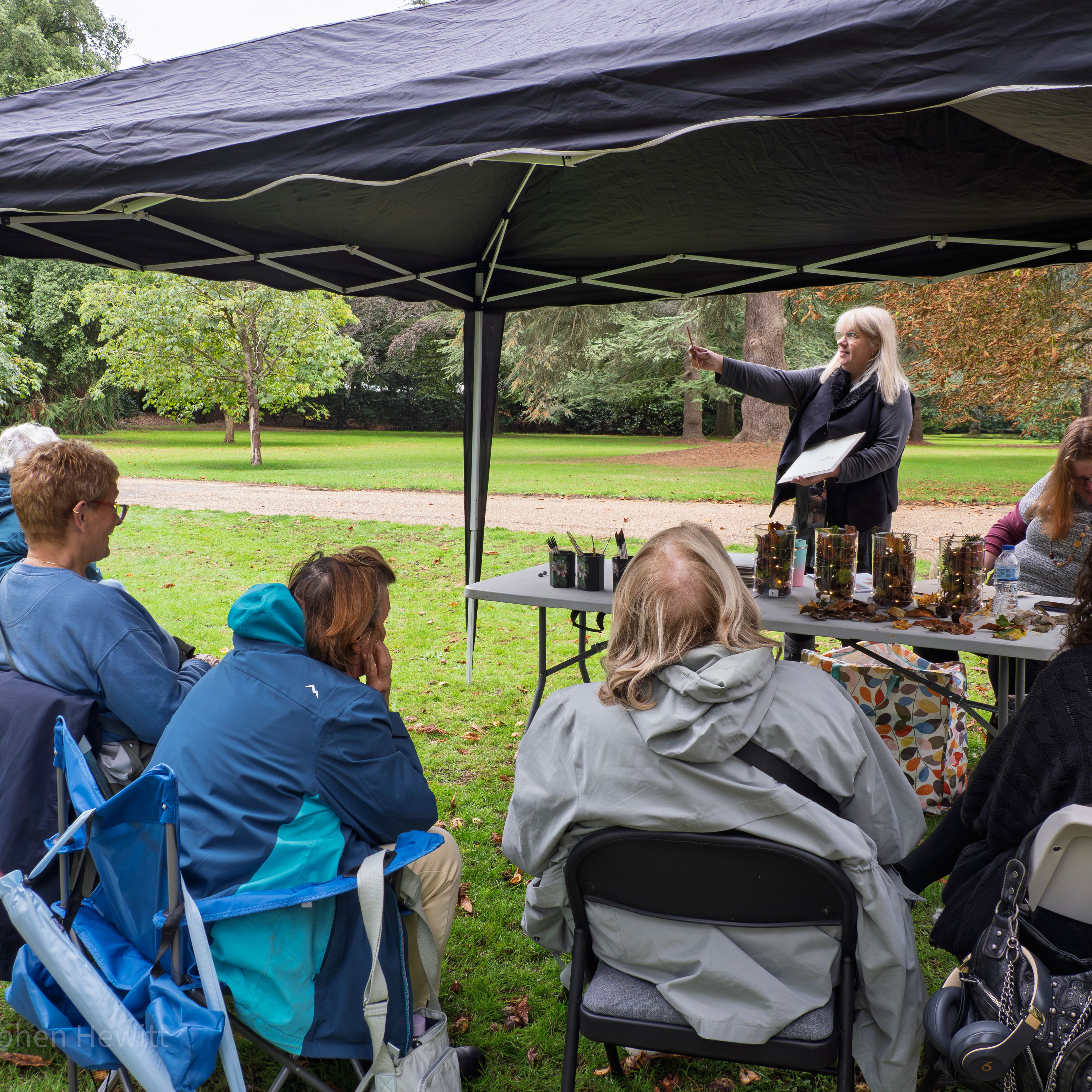 A group of people sitting under a canopy tent on a lawn, listening to a woman give a presentation.