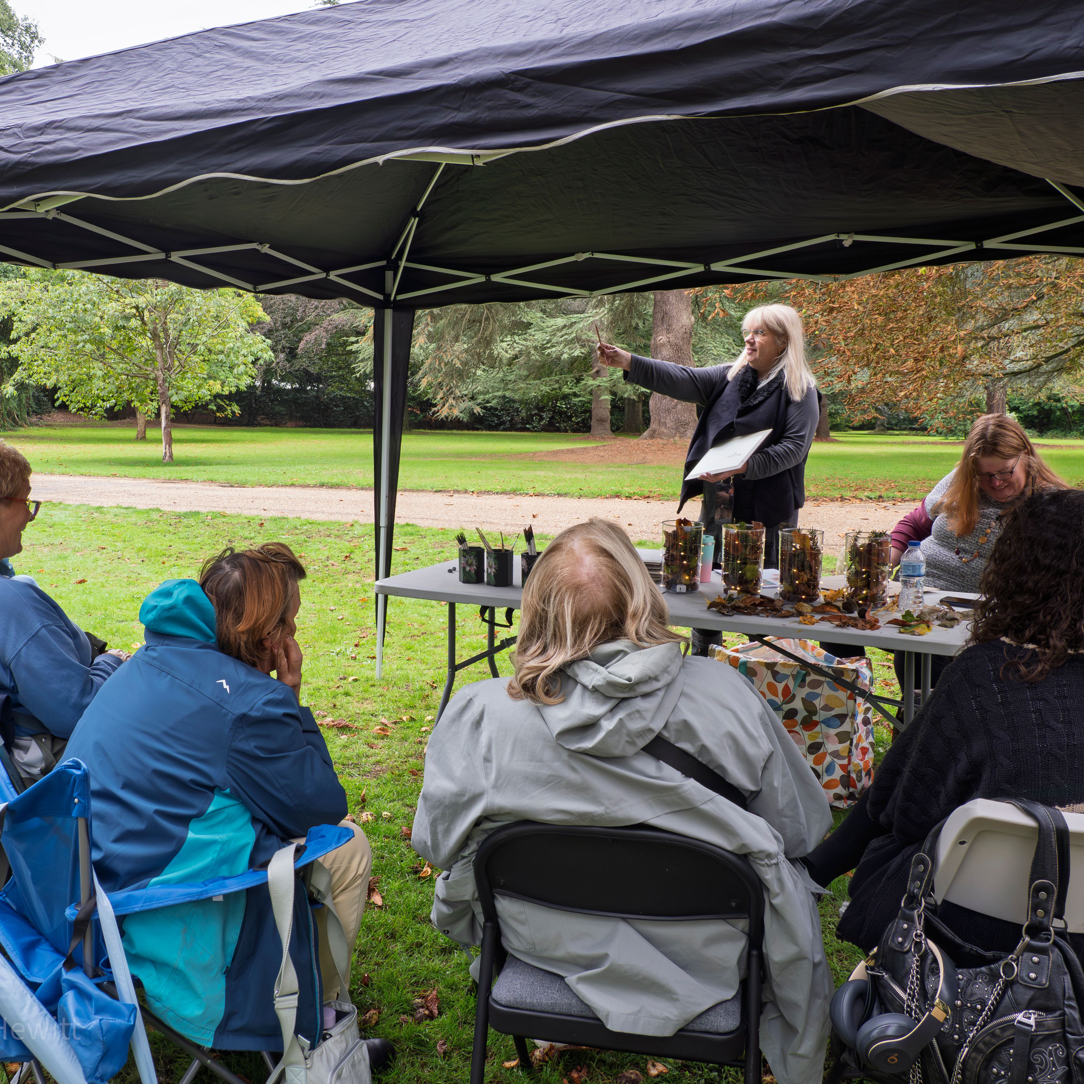 A group of people sitting under a canopy tent on a lawn, listening to a woman give a presentation.
