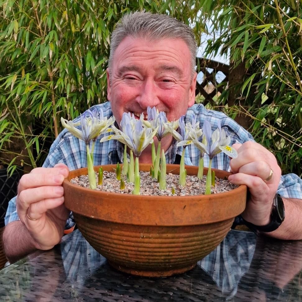 Smiling man holding a clay pot of blooming irises in a garden setting.