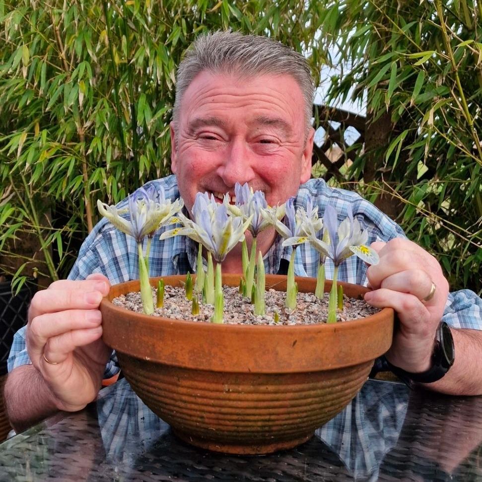 Smiling man holding a clay pot of blooming irises in a garden setting.