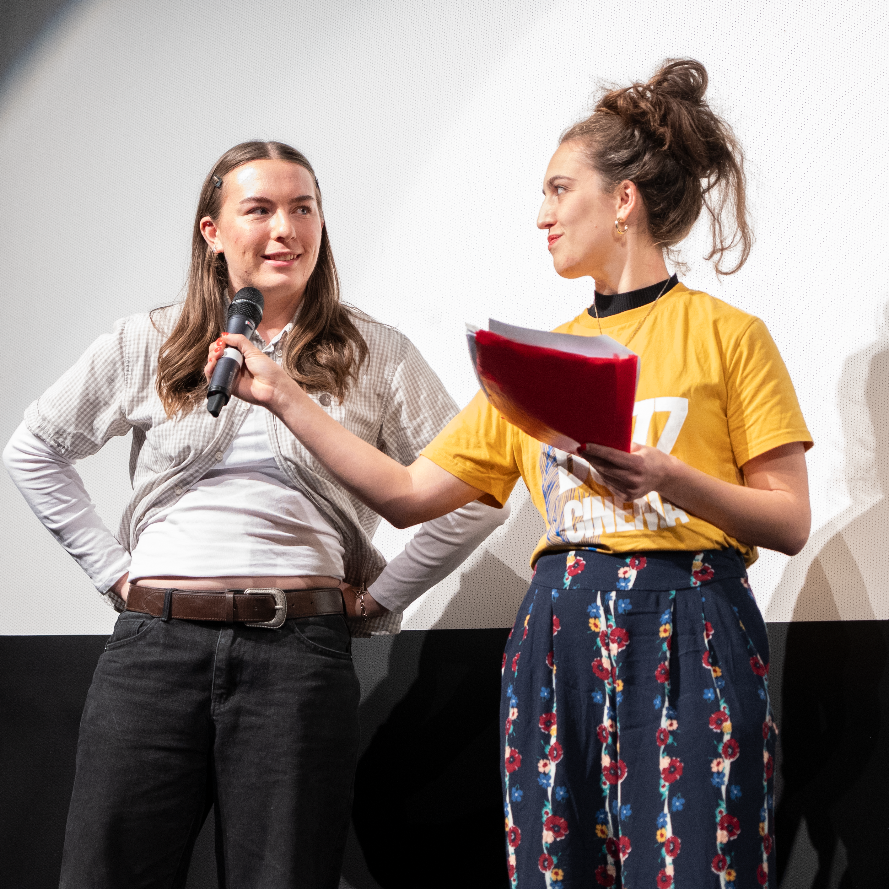 Two women standing on stage, one holding a microphone and the other holding papers