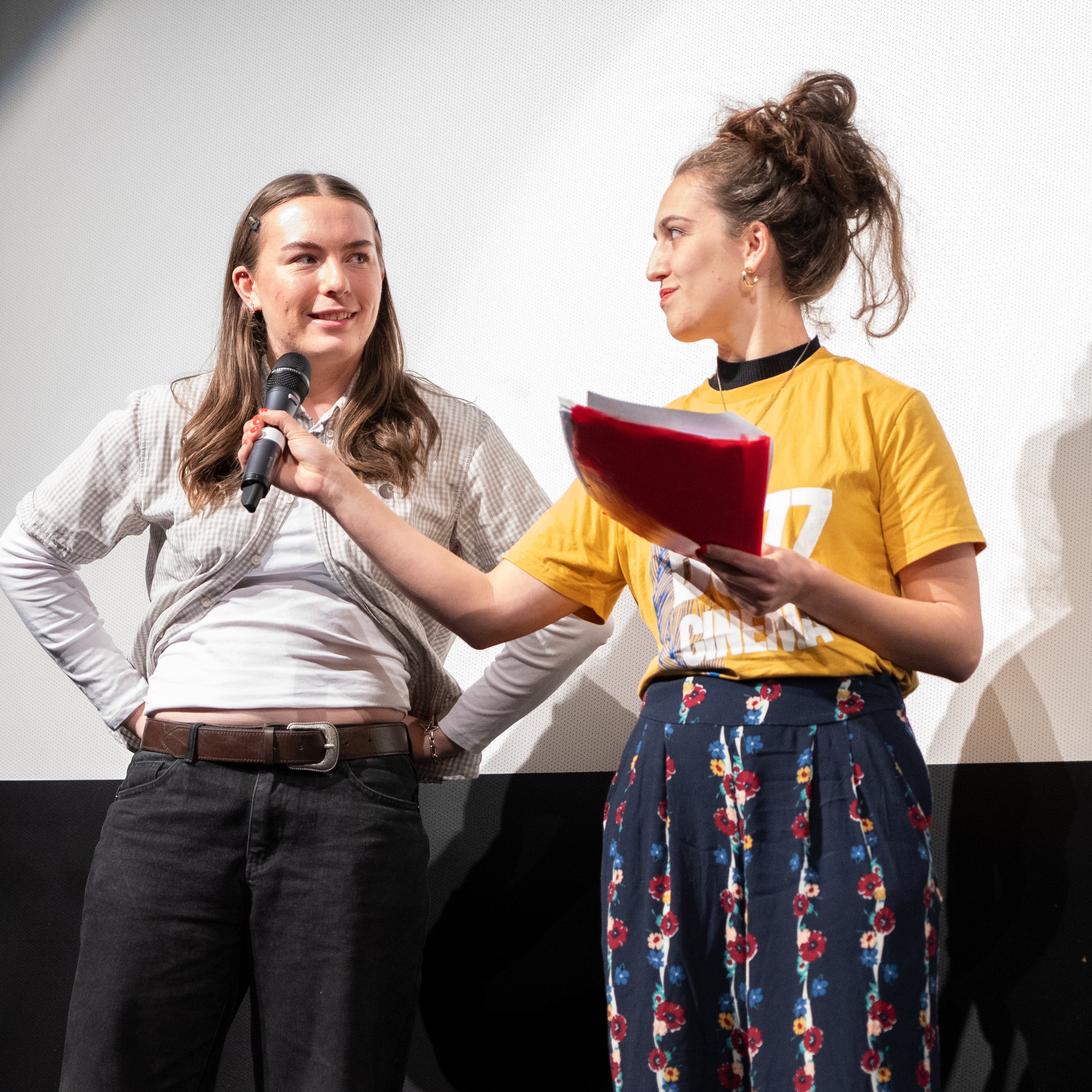 Two women standing on stage, one holding a microphone and the other holding papers