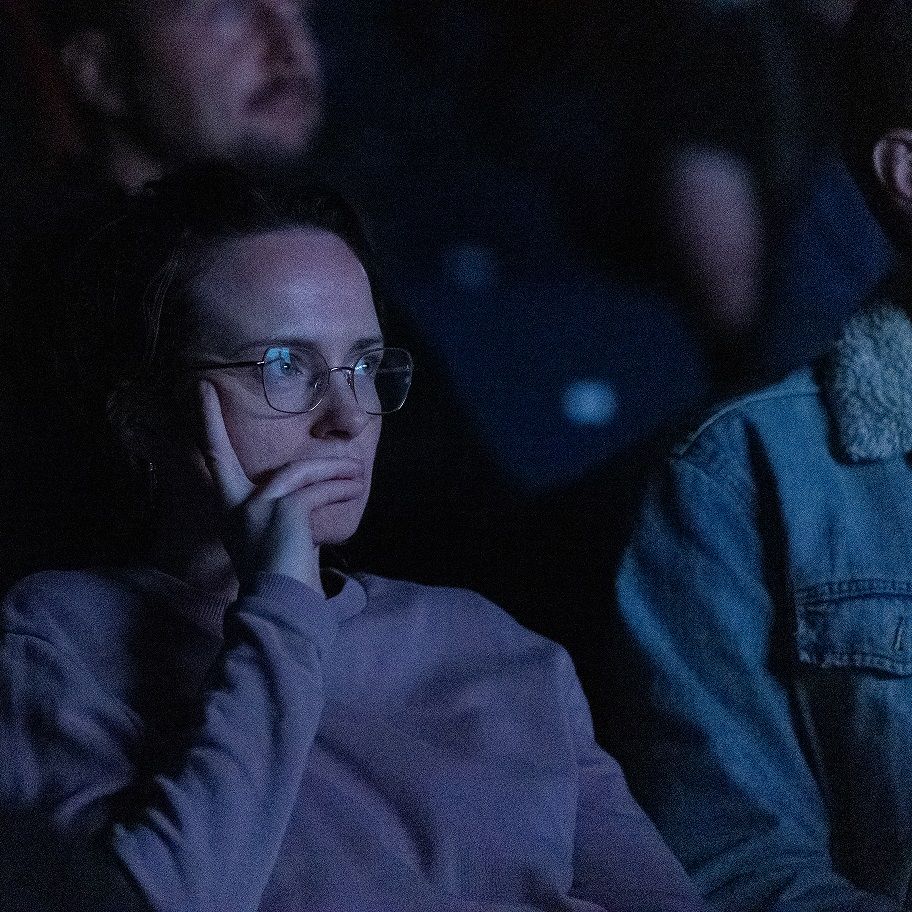 Person with glasses sitting in a dark theater, resting their face on their hand, surrounded by other people.