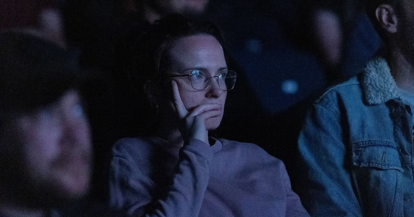 Person with glasses sitting in a dark theater, resting their face on their hand, surrounded by other people.