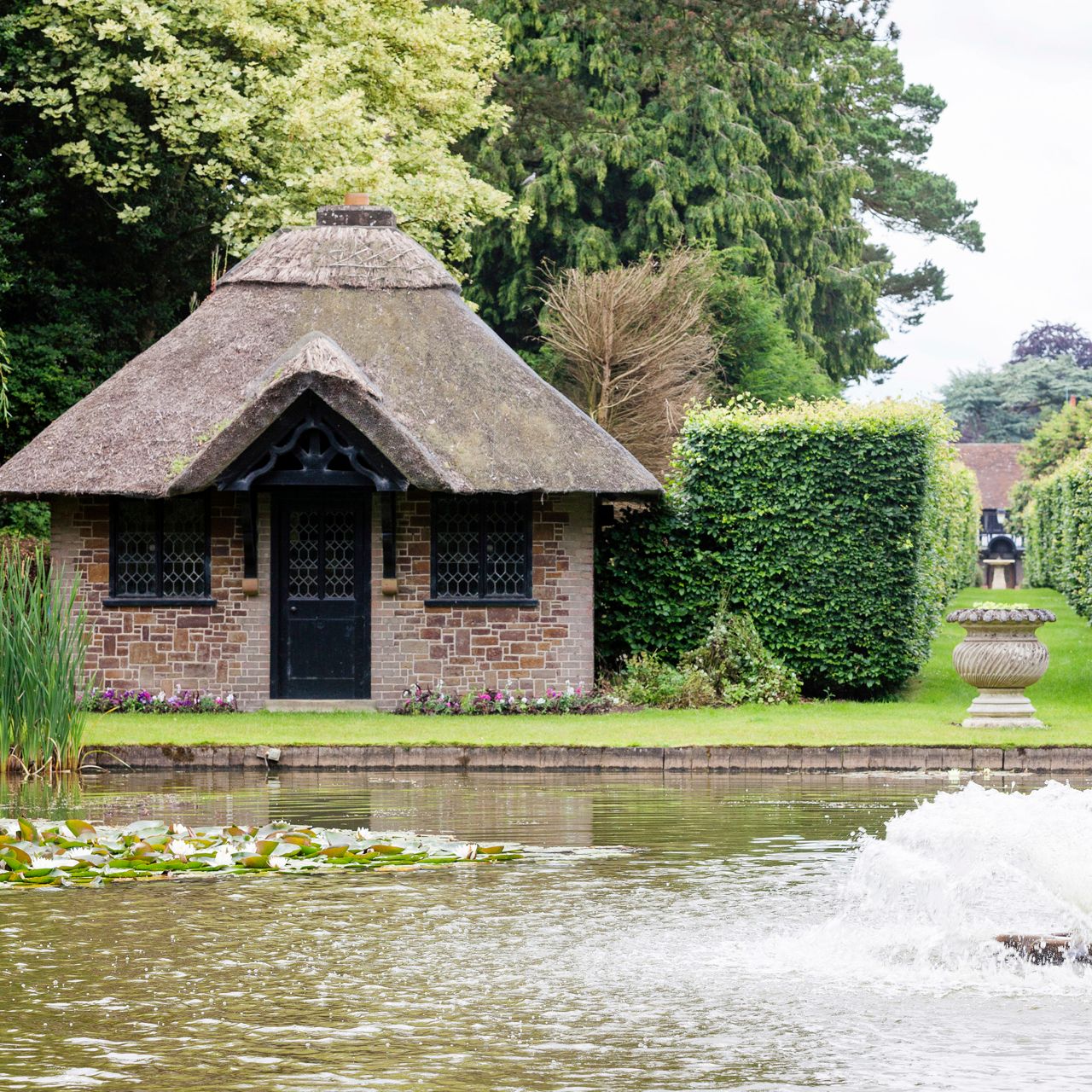 A small brick cottage with a thatched roof beside a pond with water lilies and a water fountain in the foreground.
