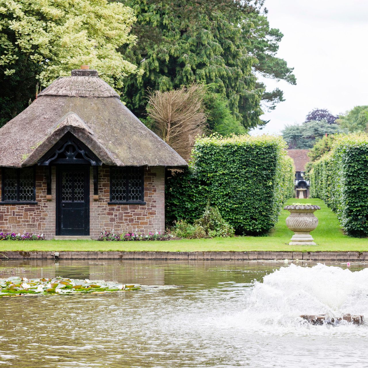 A small brick cottage with a thatched roof beside a pond with water lilies and a water fountain in the foreground.