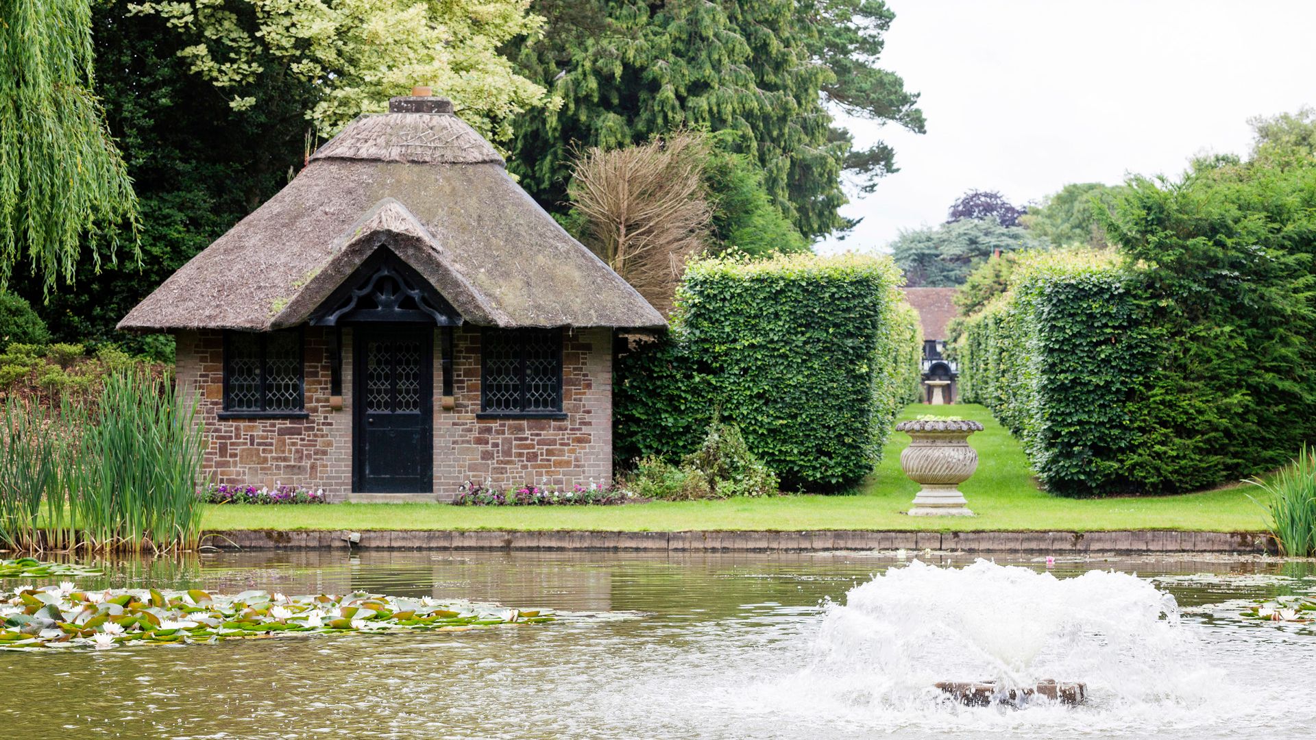 A small brick cottage with a thatched roof beside a pond with water lilies and a water fountain in the foreground.