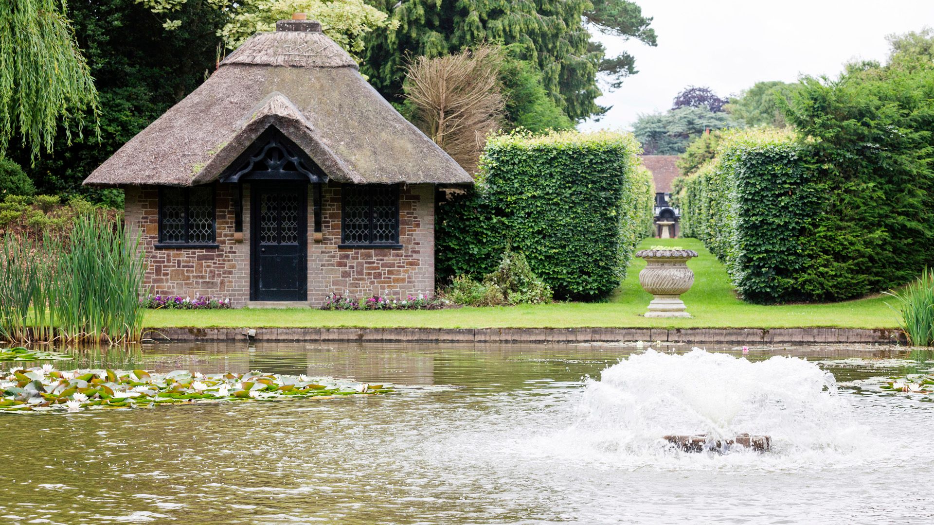 A small brick cottage with a thatched roof beside a pond with water lilies and a water fountain in the foreground.