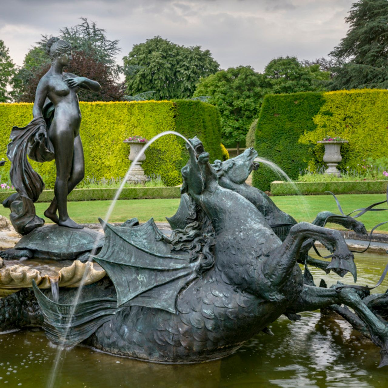 Ornate bronze fountain featuring mythological figures, sea creatures, and water sprays in a formal garden