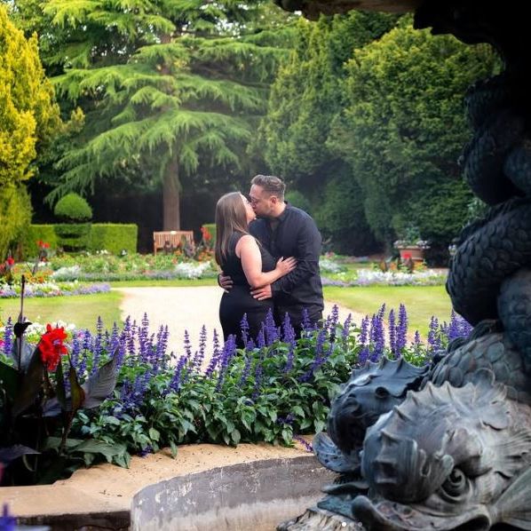 A couple kissing in a colorful garden with a large decorative fountain in the foreground.