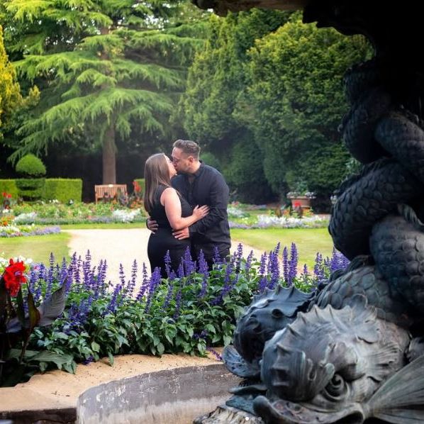 A couple kissing in a colorful garden with a large decorative fountain in the foreground.