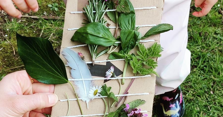 A child holding a piece of cardboard with strings, decorated with various leaves, flowers, and a feather.