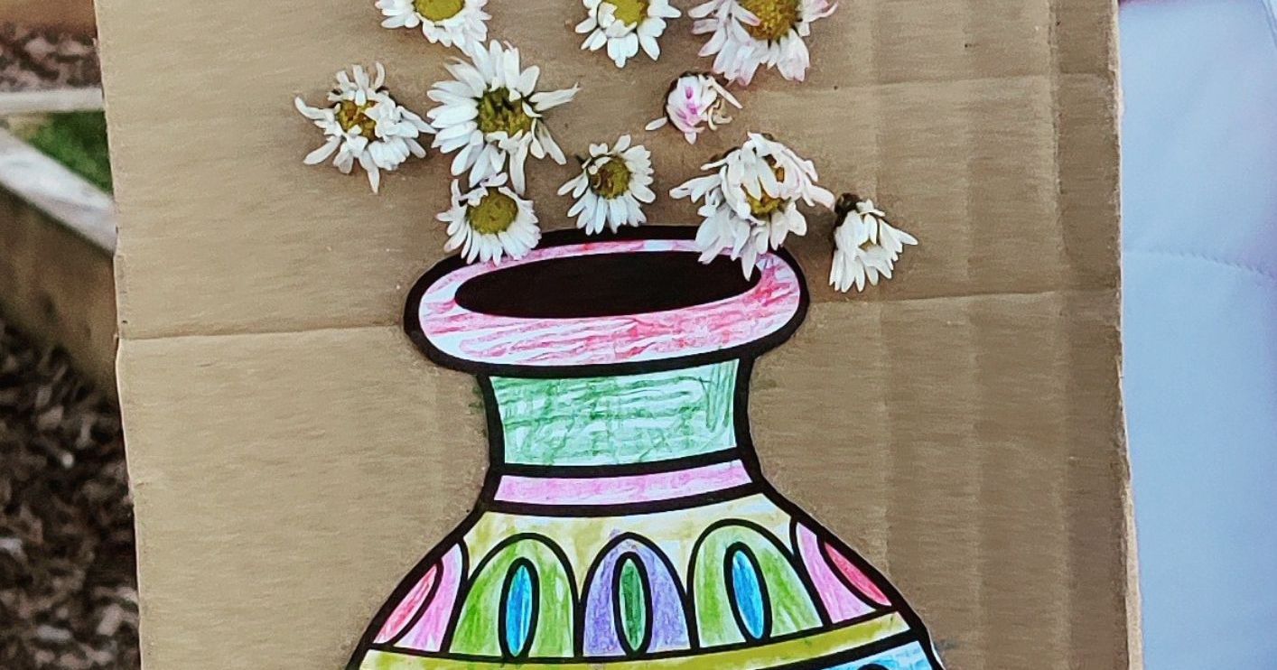 A child holds a cardboard art project showing a colored paper vase with real flowers attached above it.