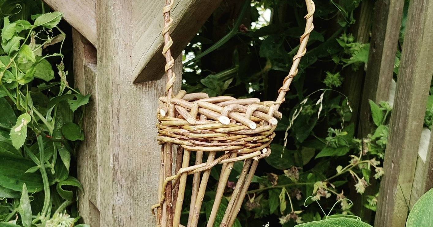 Handmade woven wicker basket hanging on a wooden structure in a garden with pink flowers and green foliage.