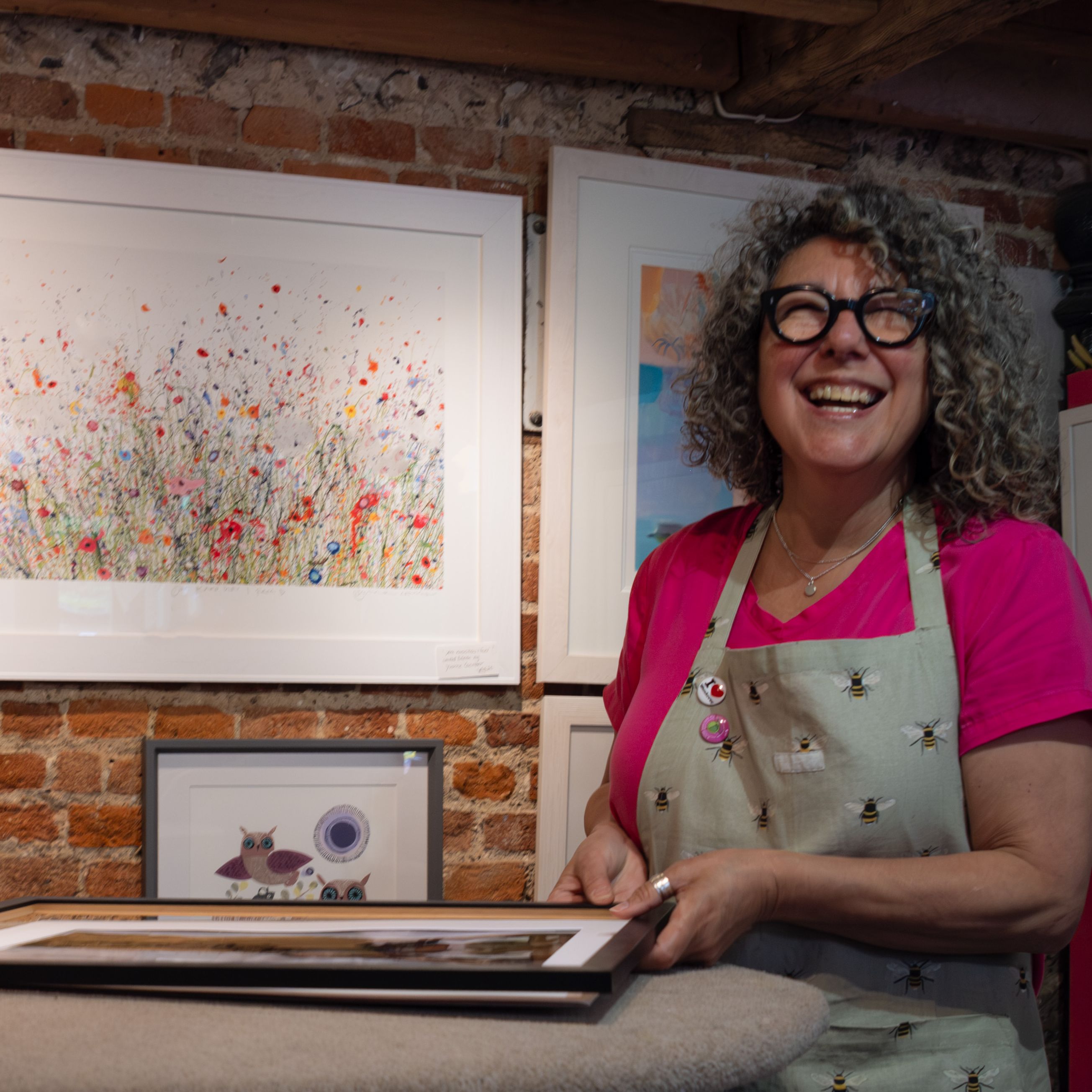 Smiling woman with curly hair and glasses standing in an art gallery, wearing an apron and holding a framed artwork.