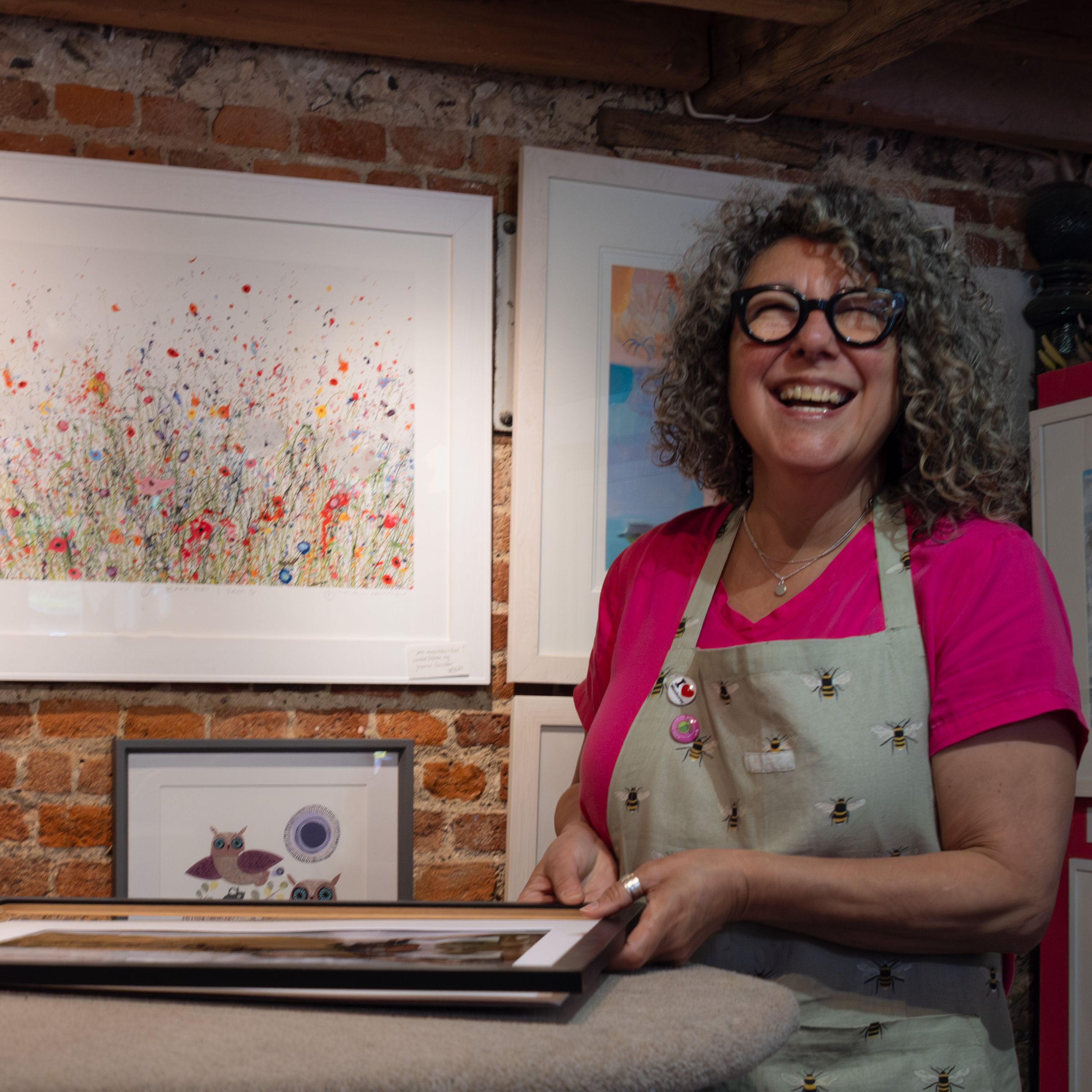 Smiling woman with curly hair and glasses standing in an art gallery, wearing an apron and holding a framed artwork.