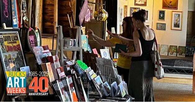 Two women browsing artwork and crafts at Bucks Art Weeks event with various art pieces and merchandise displayed.