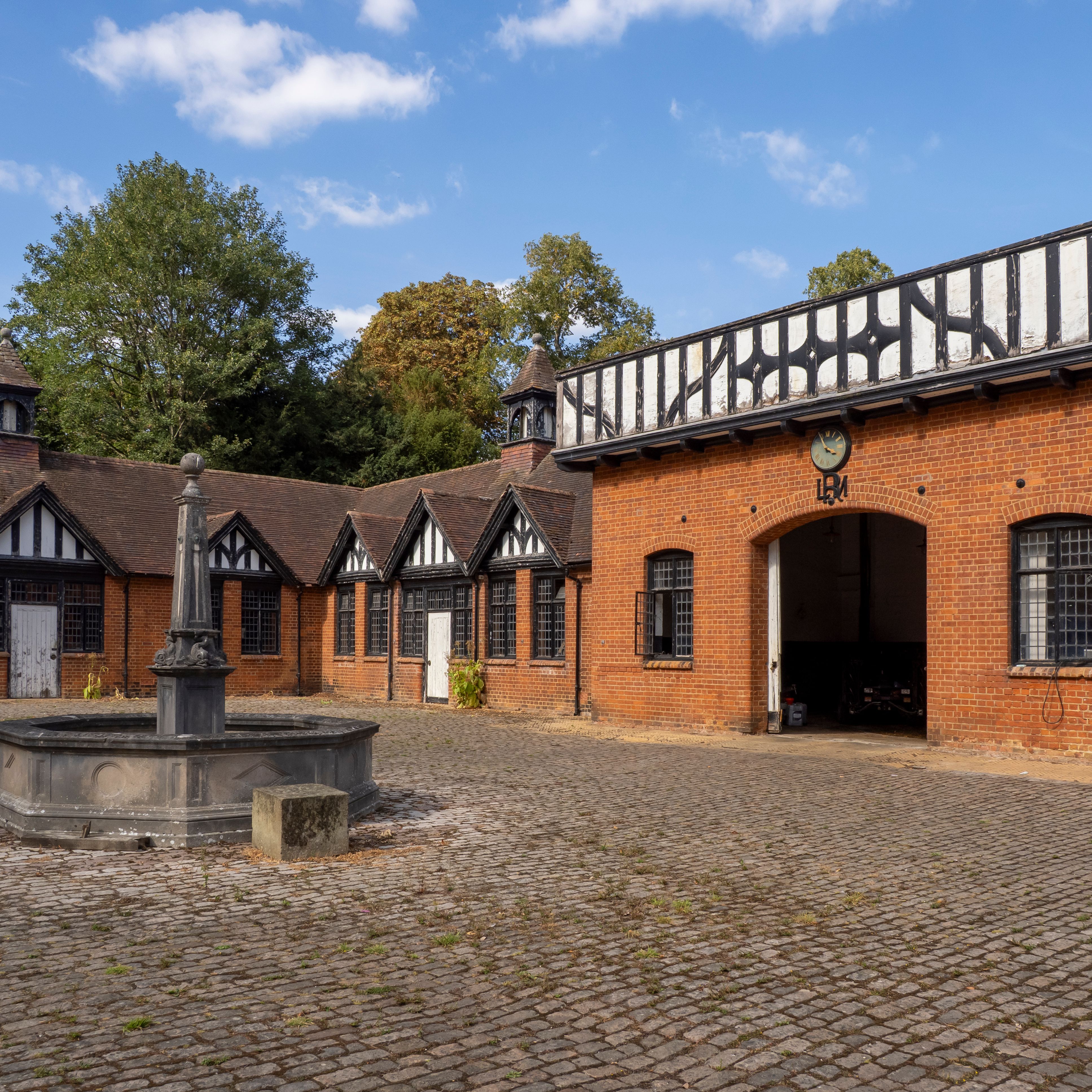 Traditional brick courtyard with a central stone fountain, surrounded by Tudor-style buildings and a clock over the main entrance.