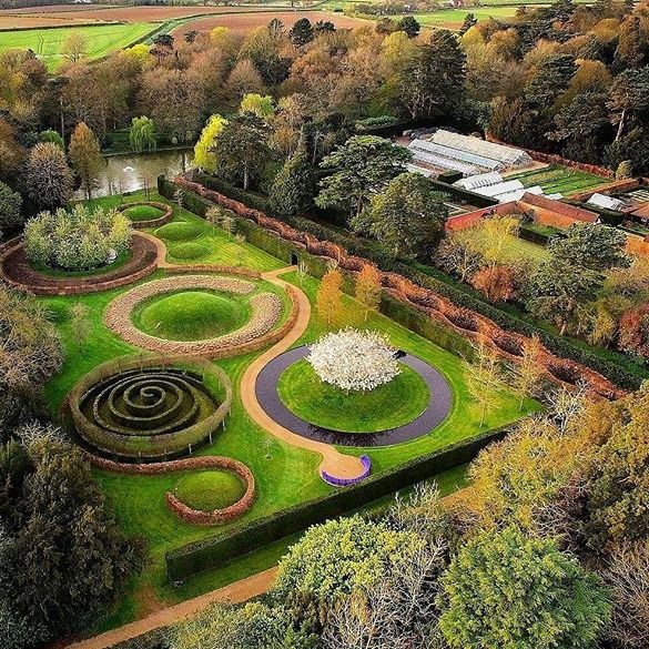 Aerial view of a formal garden with winding paths, circular and spiral hedge designs, trees, and greenhouse structures.