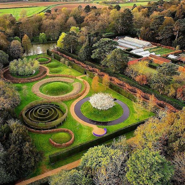 Aerial view of a formal garden with winding paths, circular and spiral hedge designs, trees, and greenhouse structures.