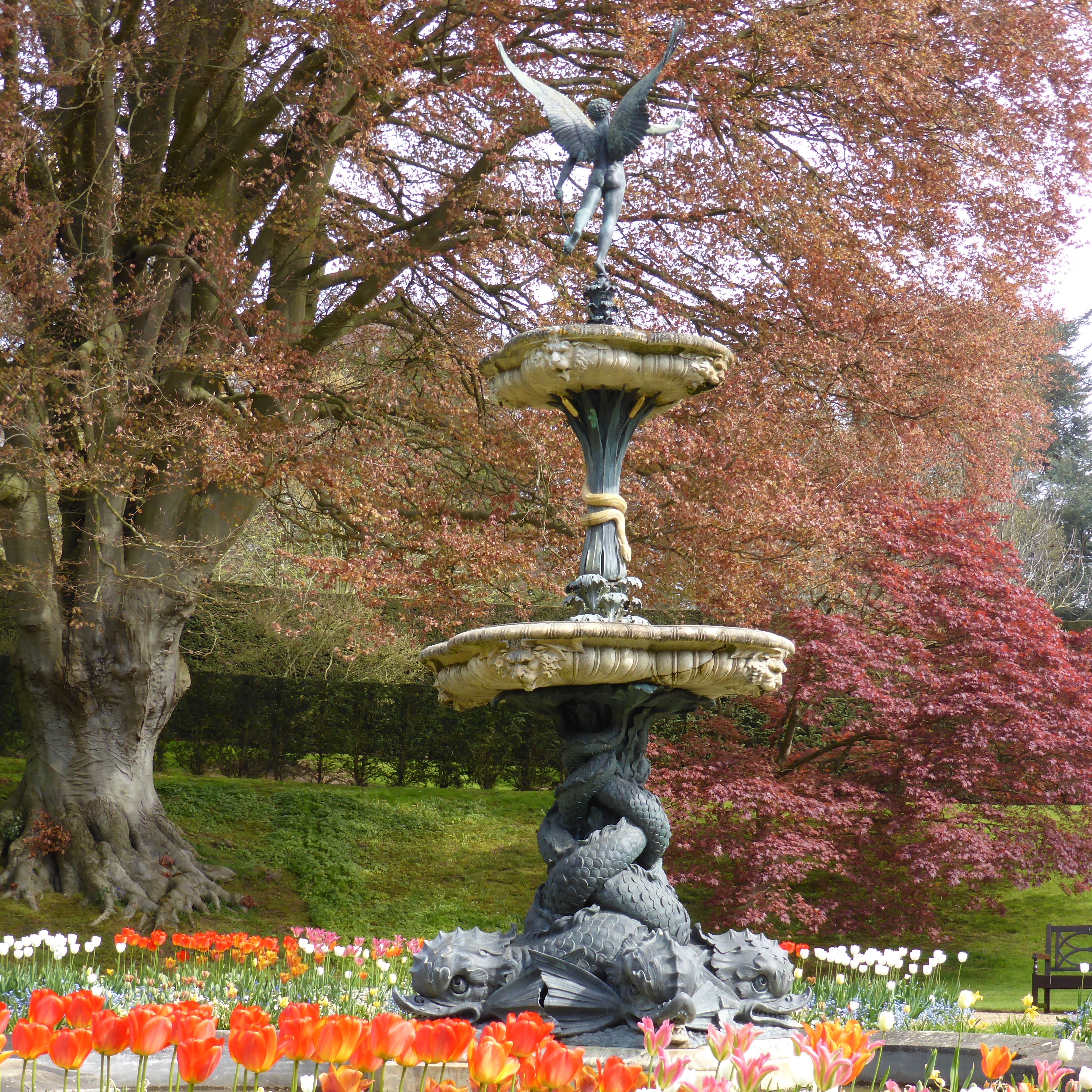Decorative fountain with an angel statue, surrounded by colorful spring flowers and trees with reddish leaves.