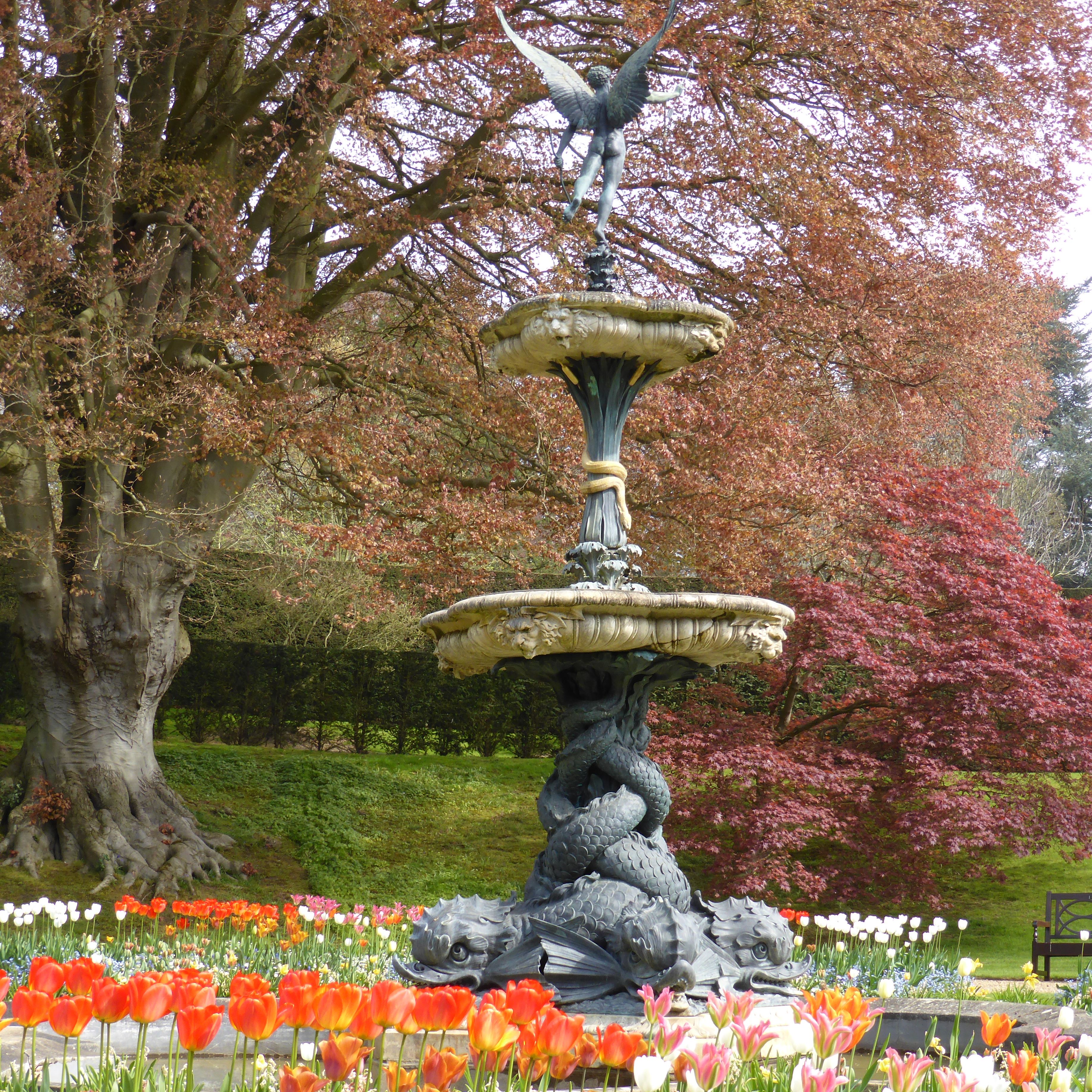 Decorative fountain with an angel statue, surrounded by colorful spring flowers and trees with reddish leaves.