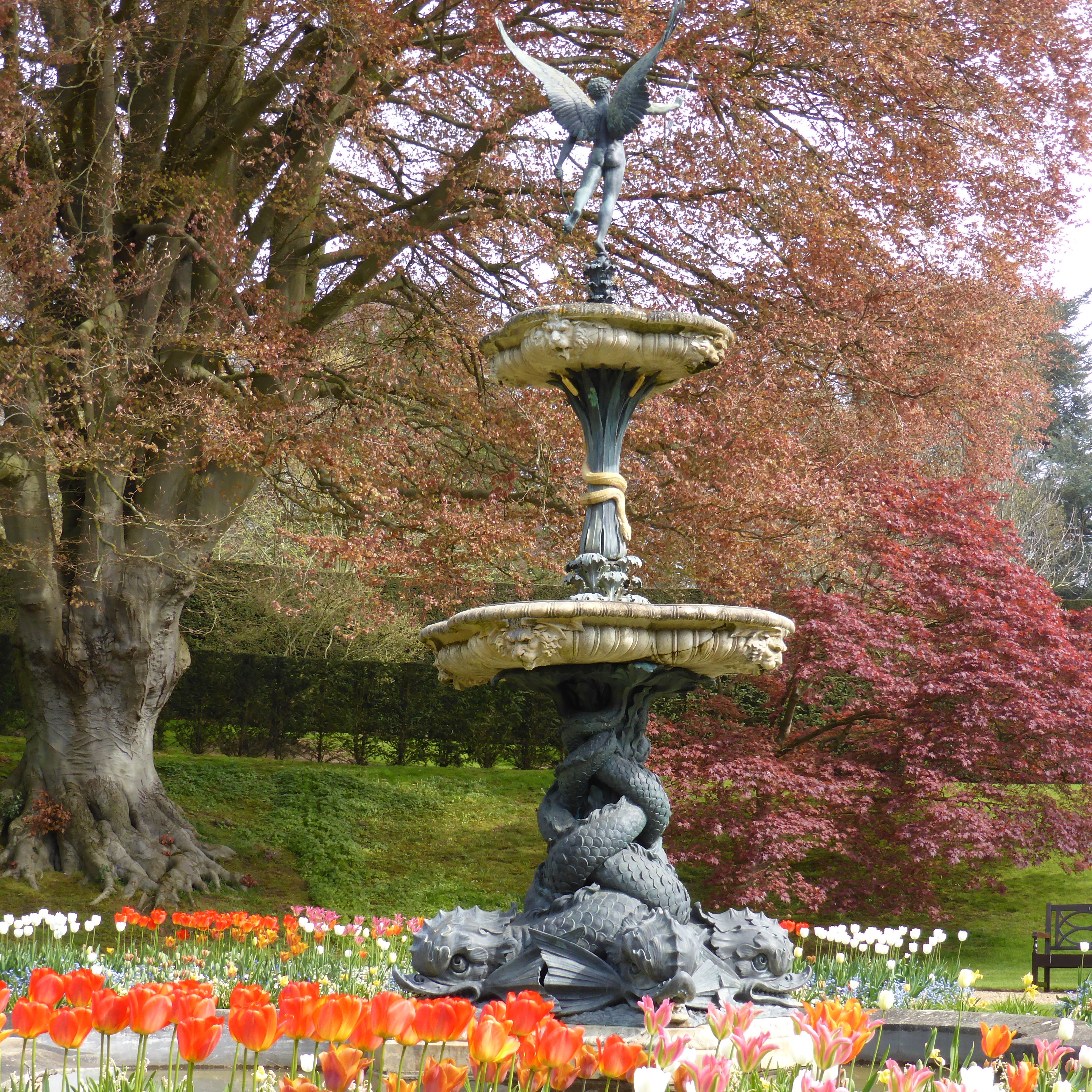 Decorative fountain with an angel statue, surrounded by colorful spring flowers and trees with reddish leaves.