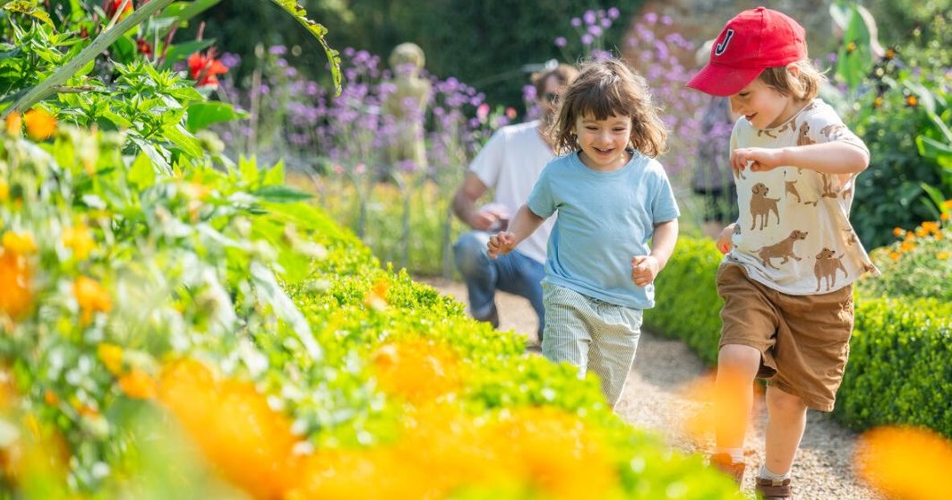 Two children running along a garden path bordered by colourful flowers, with an adult in the background