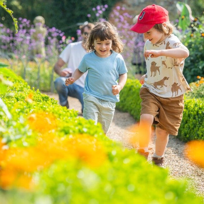 Two children running along a garden path bordered by colourful flowers, with an adult in the background