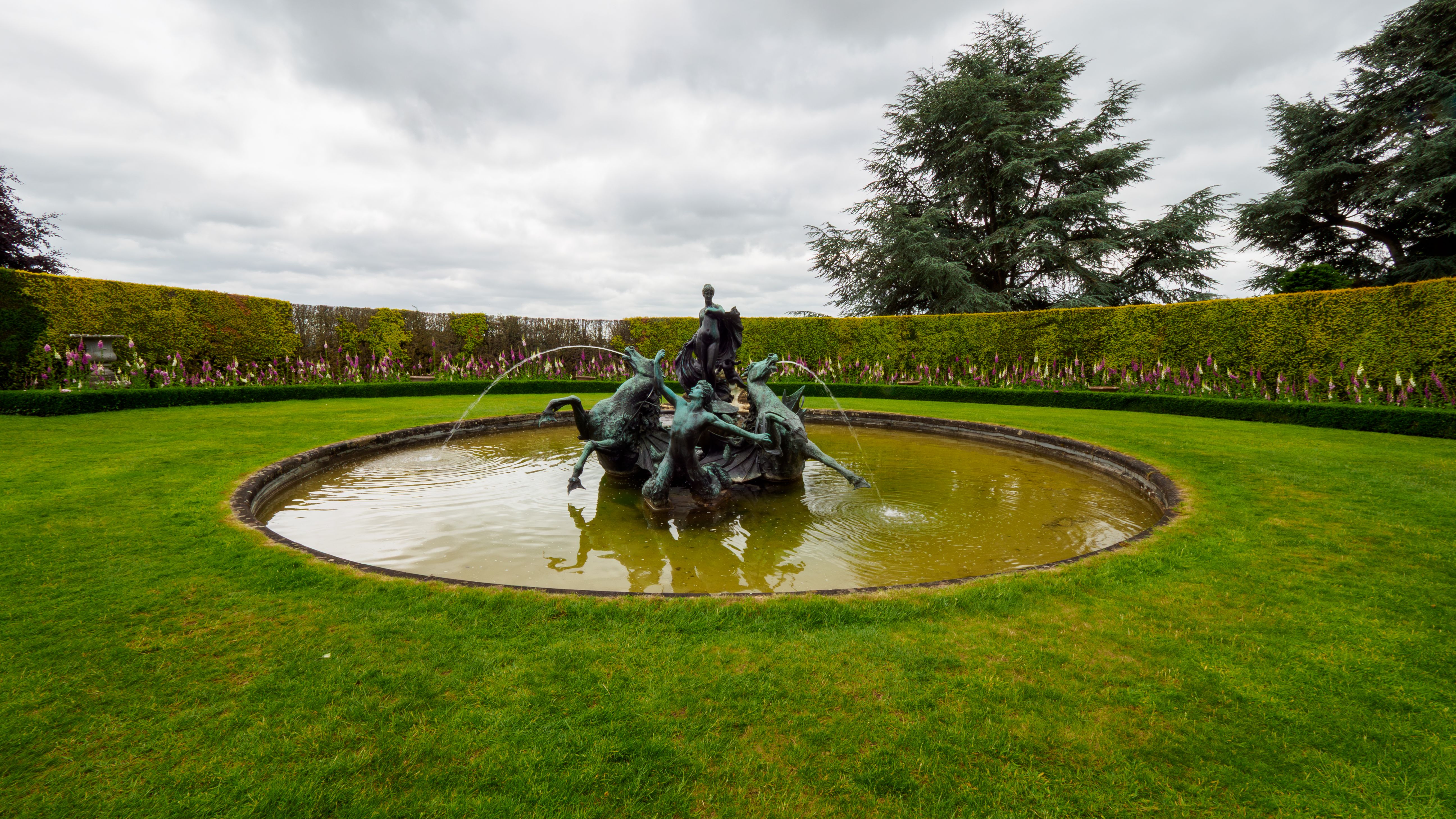 Bronze fountain sculpture in a circular pond within a formal garden, with clipped hedges and a large evergreen tree under a cloudy sky.