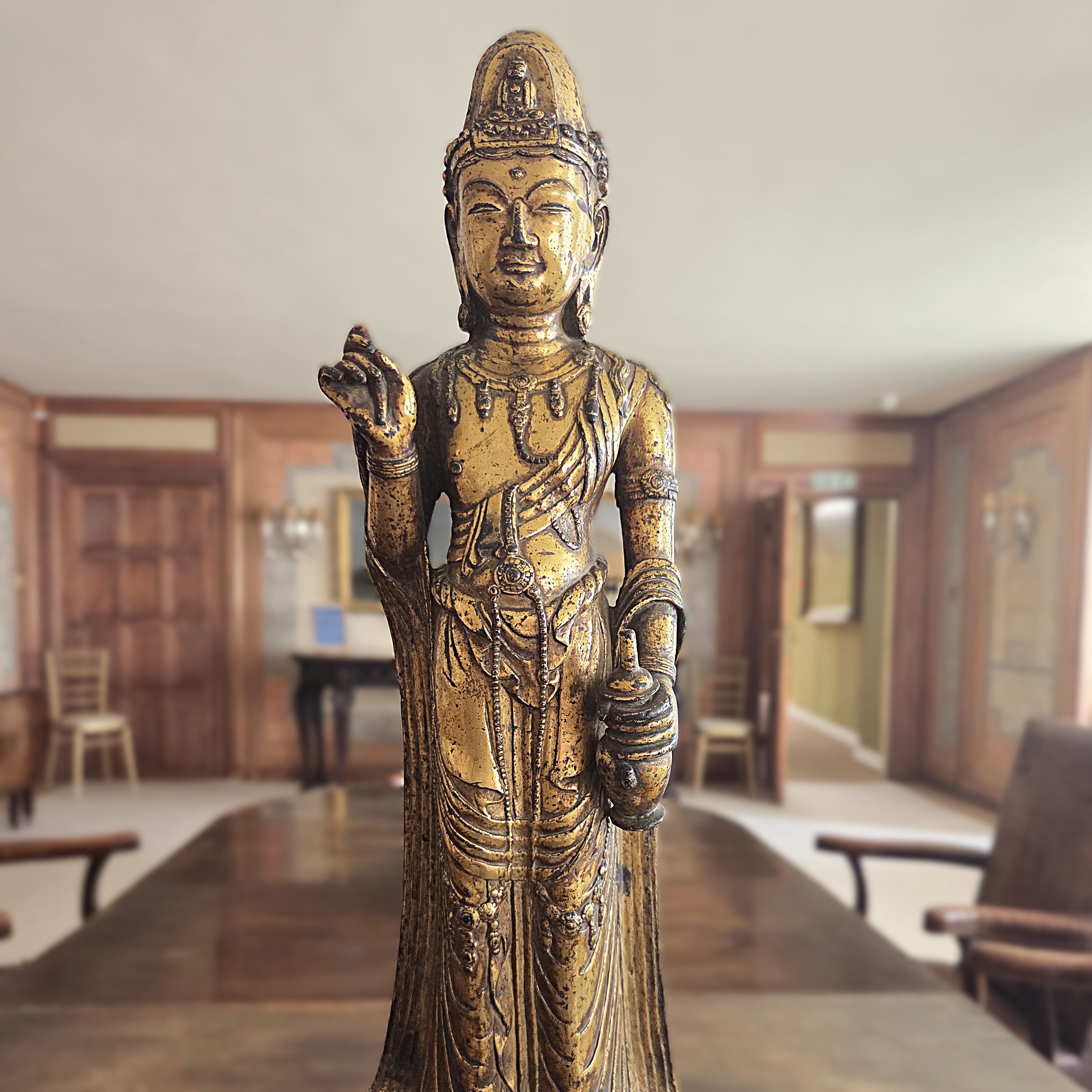 Gold-coloured statue of a deity holding a vessel, displayed indoors on a wooden base