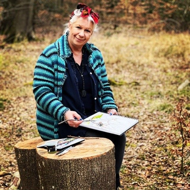Woman sitting on a tree stump in a woodland, sketching on a pad with art supplies beside her