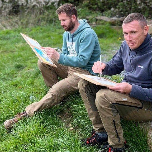 Two men sitting on grass outdoors, painting on drawing boards