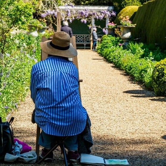 Artist seated on a folding stool sketching in a sunlit garden path lined with flowers and wisteria