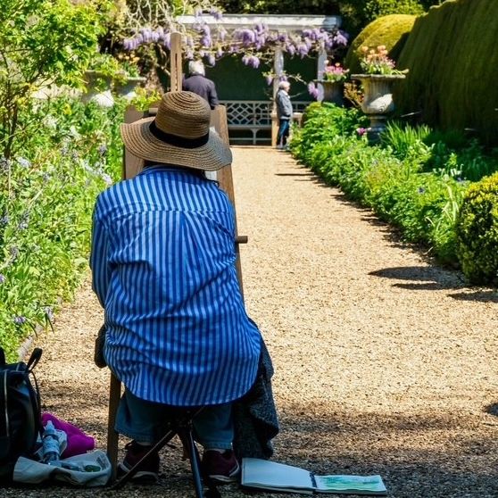 Artist seated on a folding stool sketching in a sunlit garden path lined with flowers and wisteria