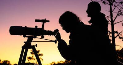 Silhouettes of two people stargazing with a telescope at twilight
