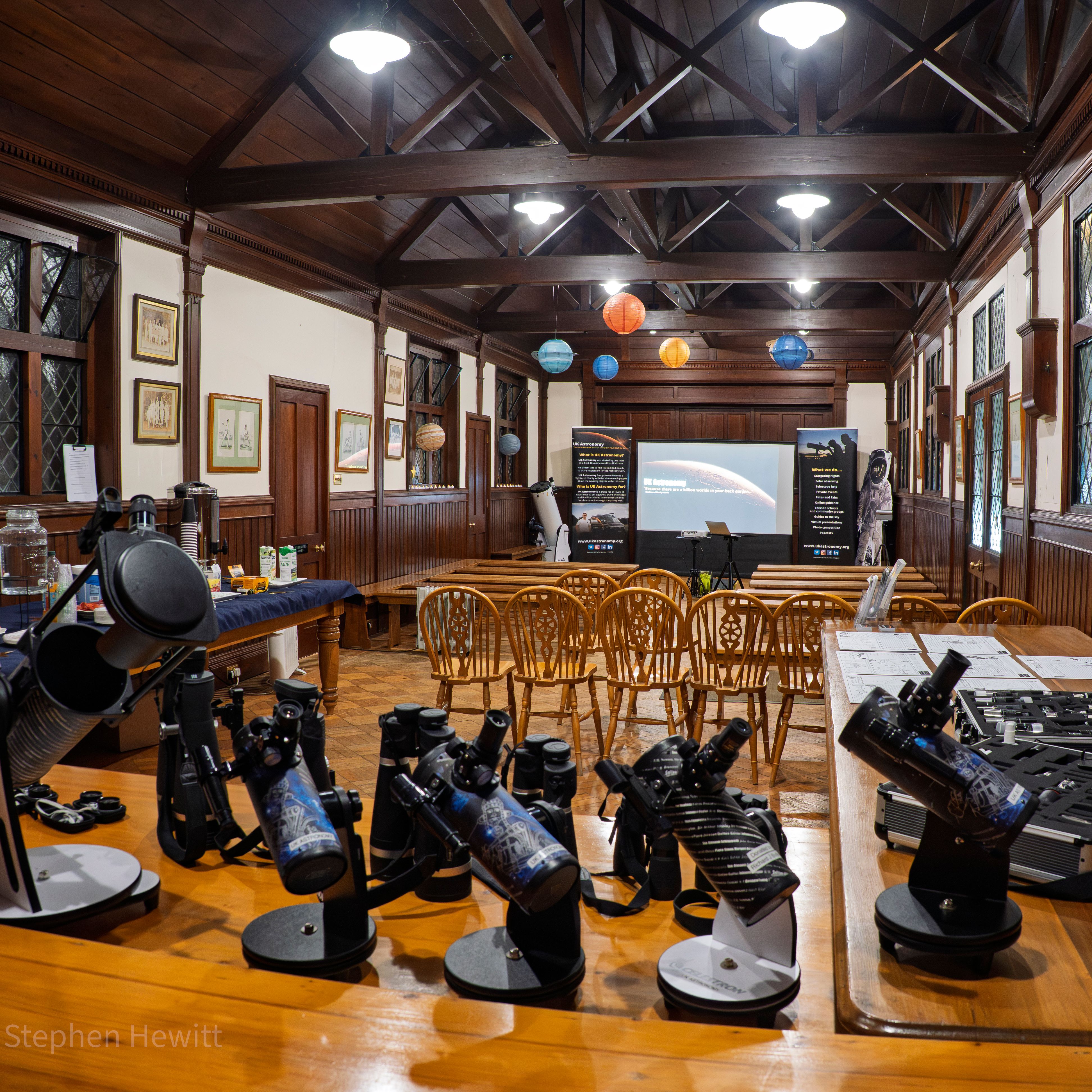 Wood-panelled hall set up for an astronomy workshop with telescopes on tables, chairs facing a projector screen, and hanging paper planets