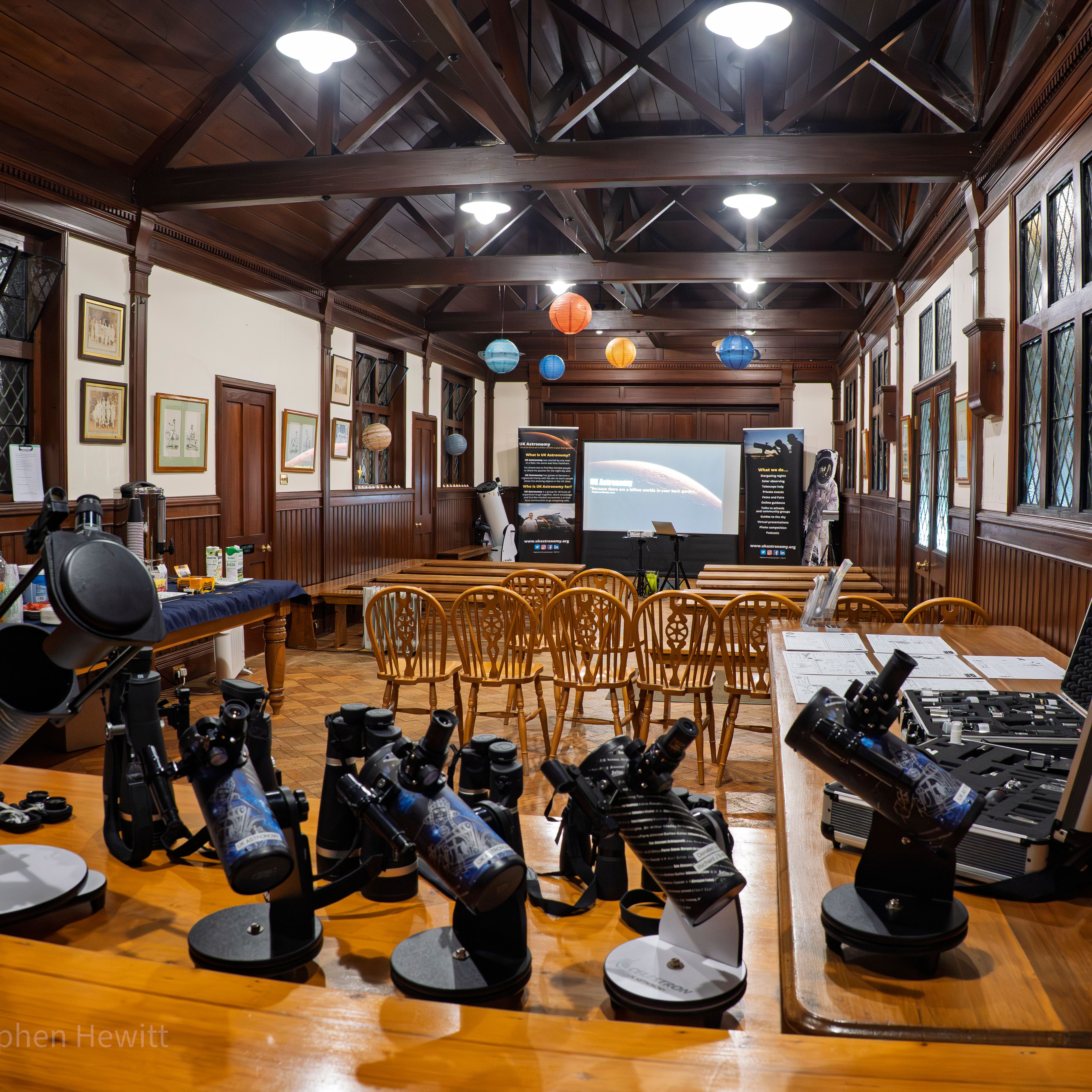Wood-panelled hall set up for an astronomy workshop with telescopes on tables, chairs facing a projector screen, and hanging paper planets