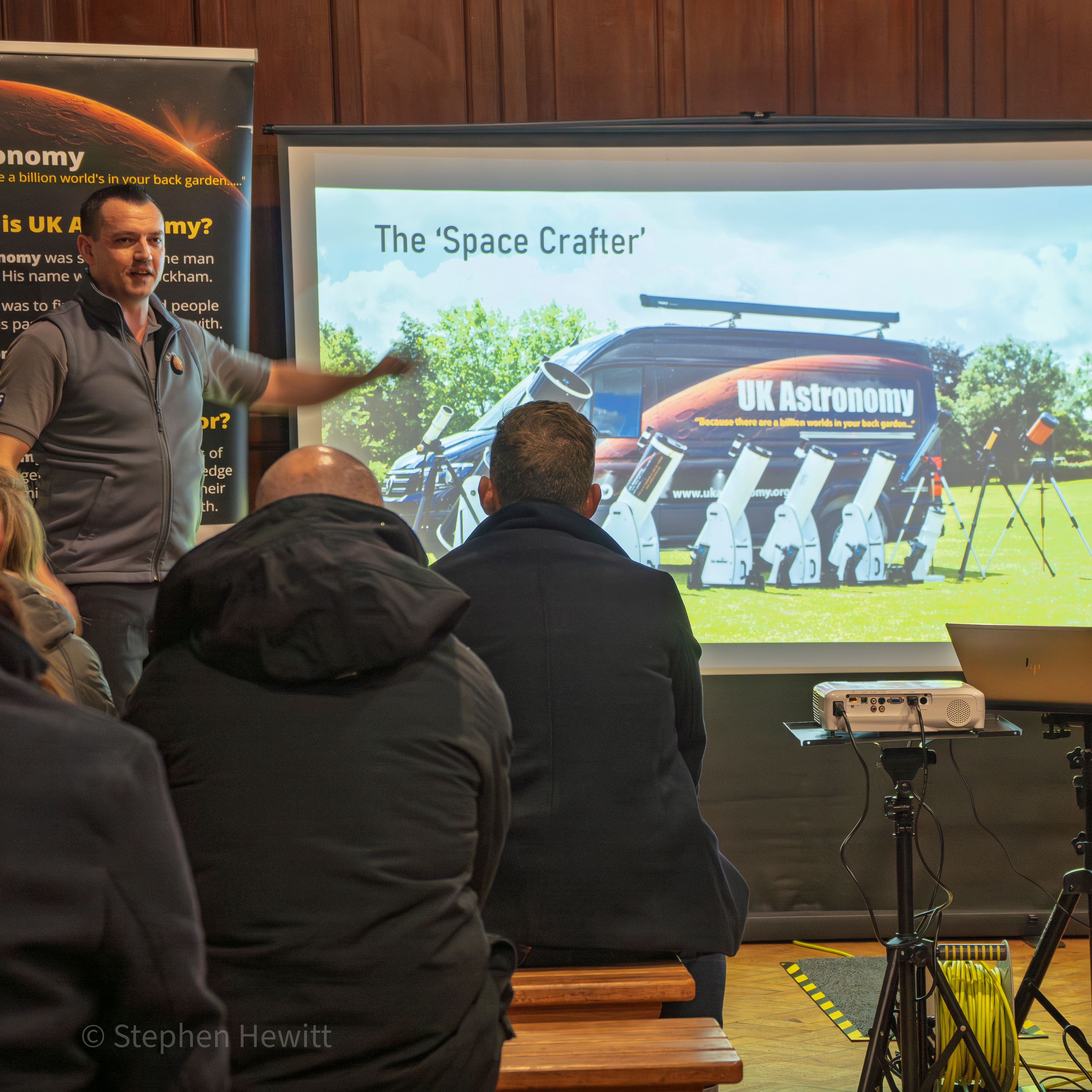 Presenter speaking to an audience beside a projected slide titled 'The Space Crafter', showing a UK Astronomy van and telescopes.