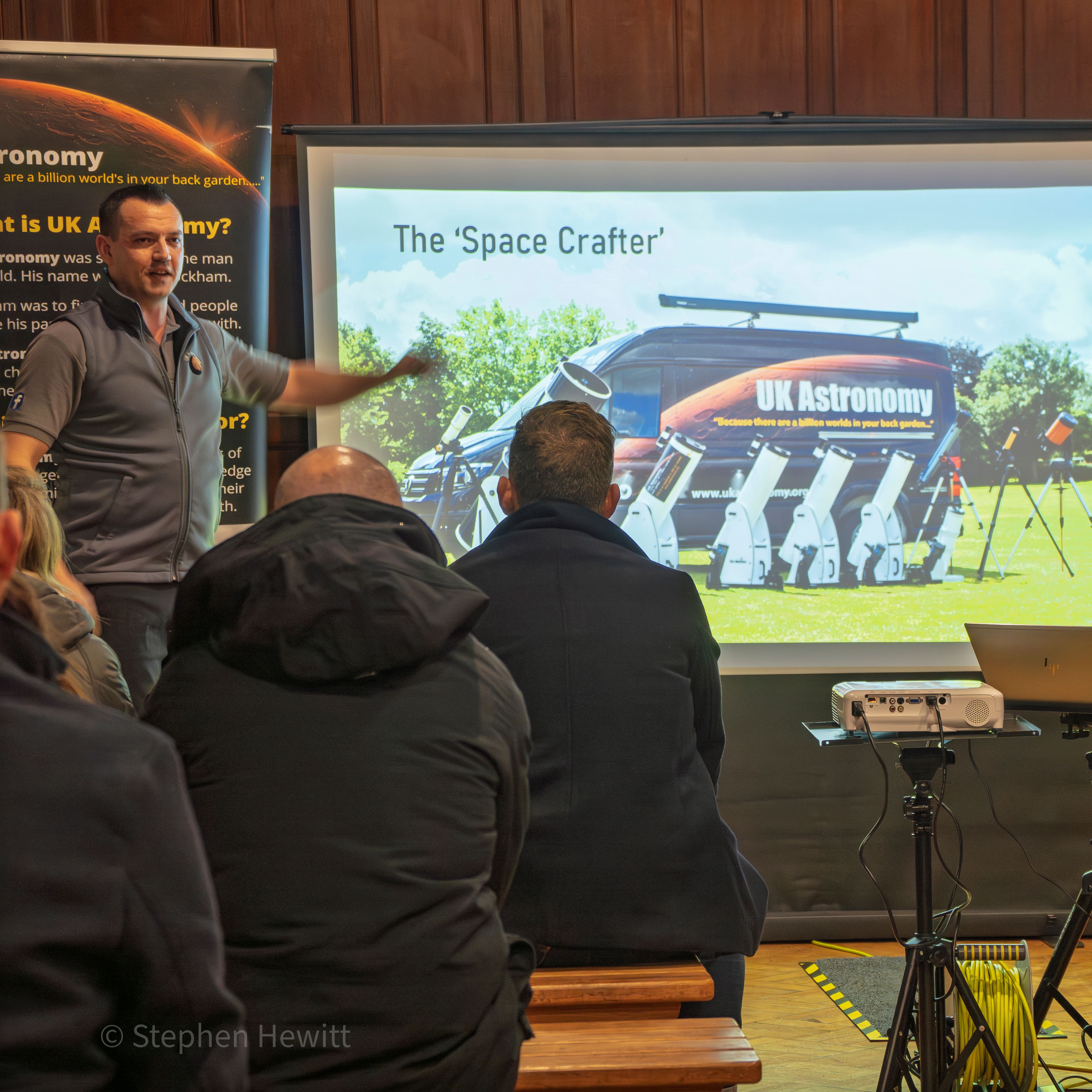 Presenter speaking to an audience beside a projected slide titled 'The Space Crafter', showing a UK Astronomy van and telescopes.