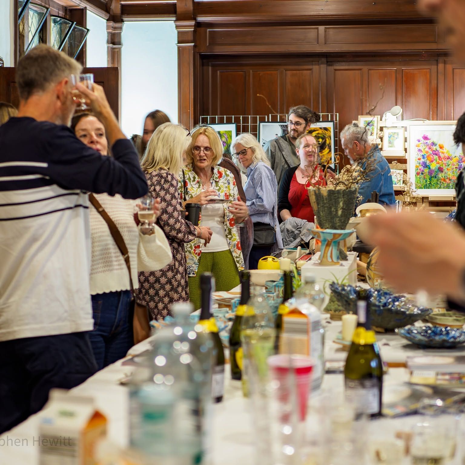 People chatting at an indoor art exhibition with colourful paintings and craft items on display