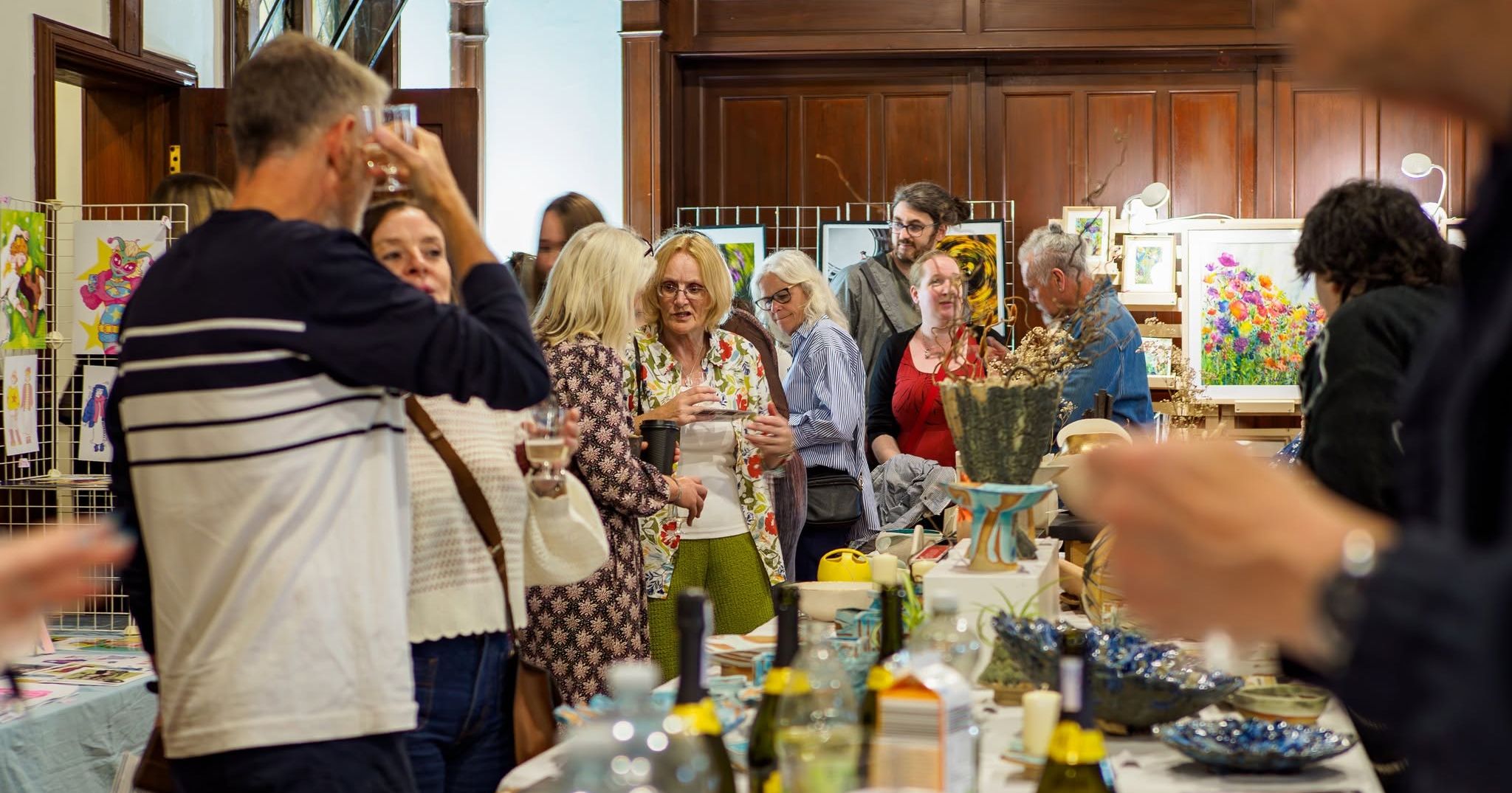 People chatting at an indoor art exhibition with colourful paintings and craft items on display