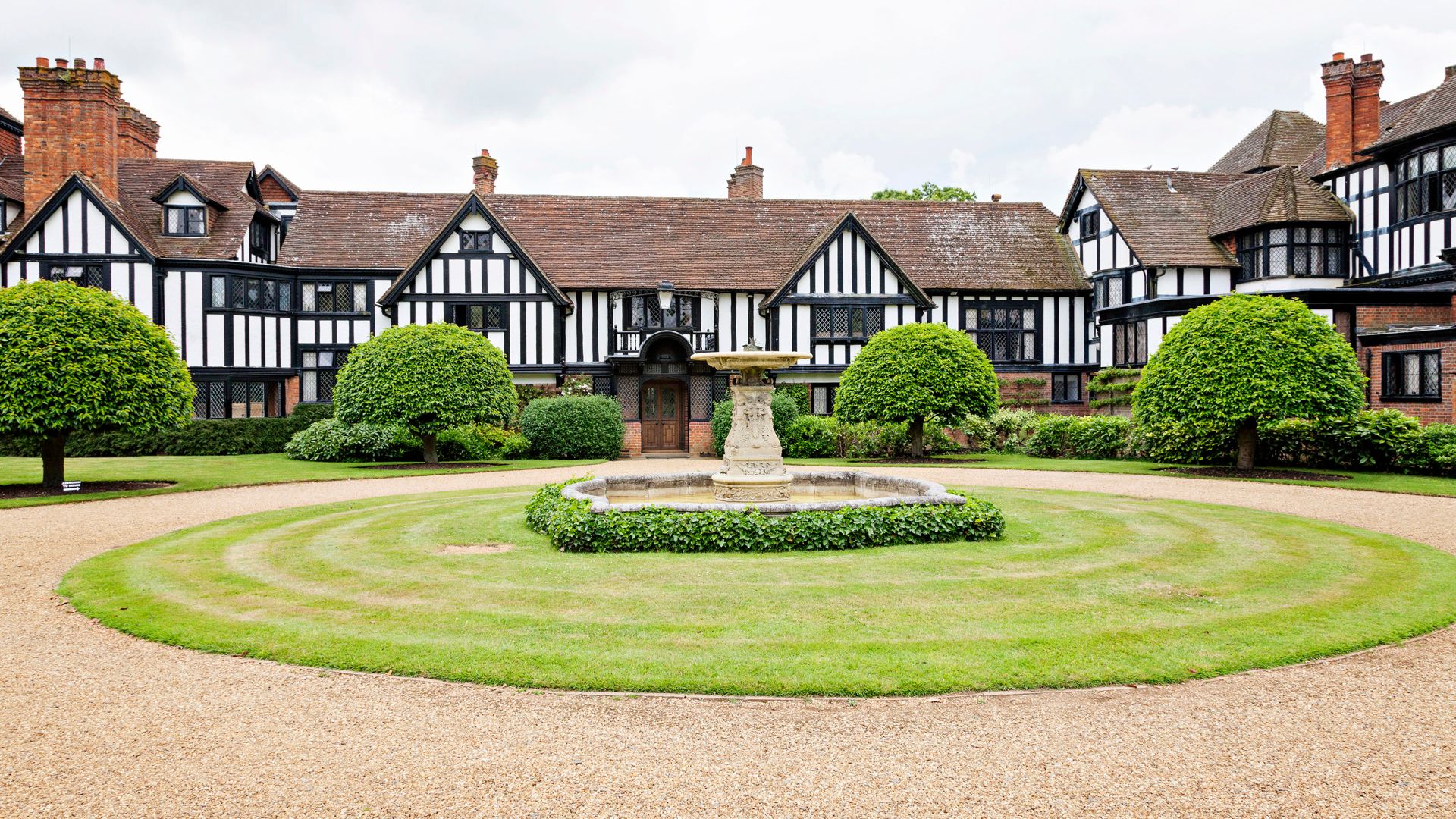Tudor-style manor house with black and white timber framing, surrounded by manicured gardens and a central stone fountain.