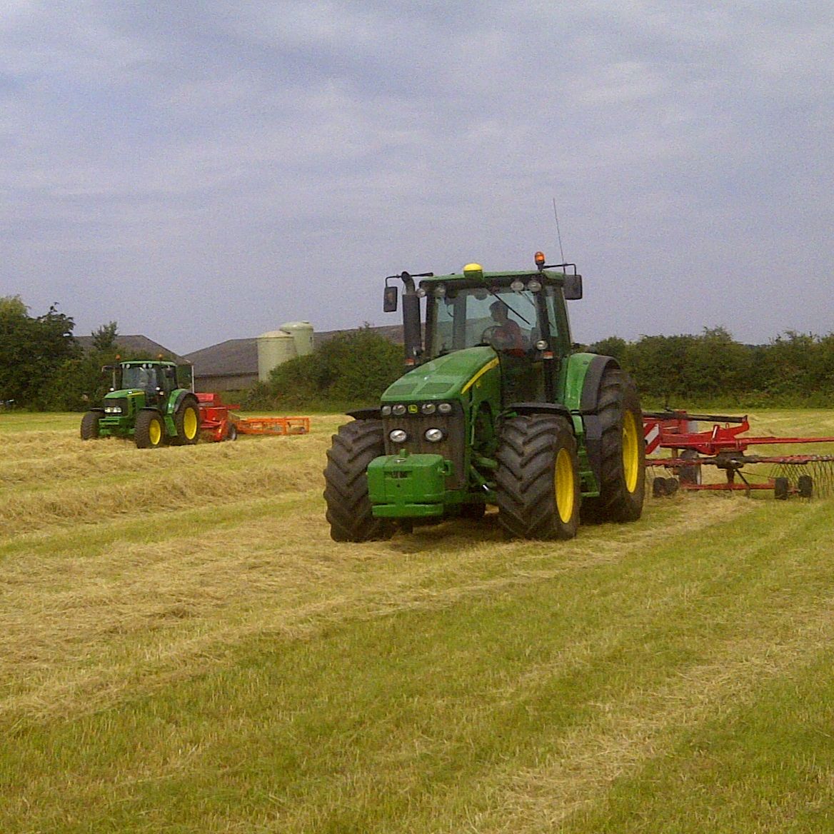 Two green tractors working in a field with cut hay.