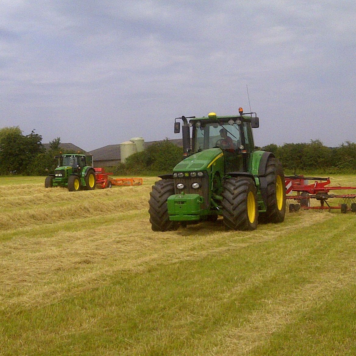 Two green tractors working in a field with cut hay.