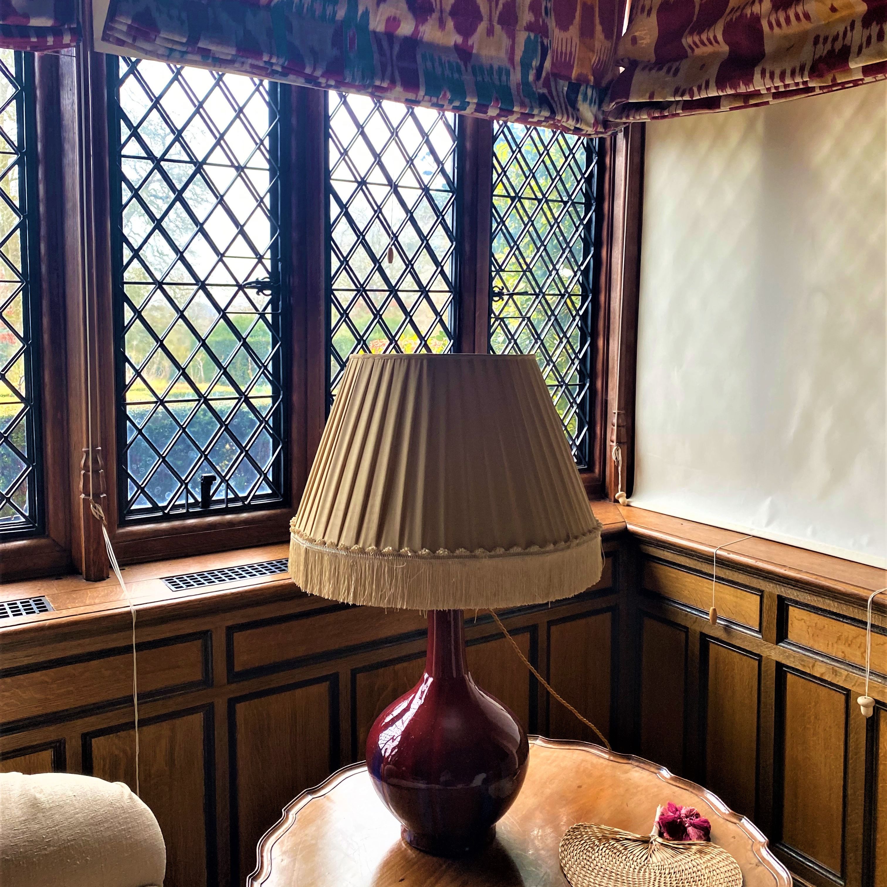 Table lamp with pleated shade on a wooden table in front of leaded glass windows.