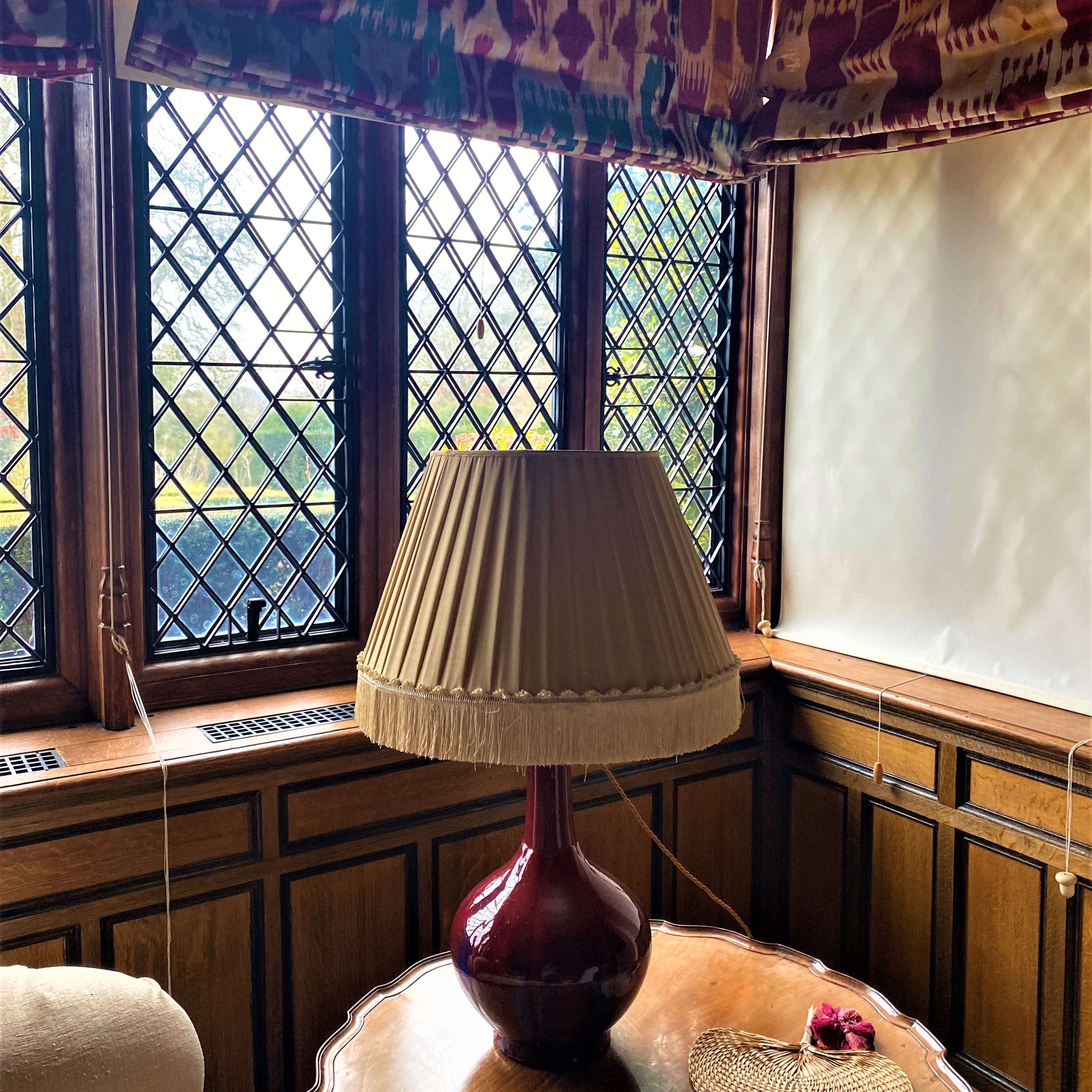 Table lamp with pleated shade on a wooden table in front of leaded glass windows.