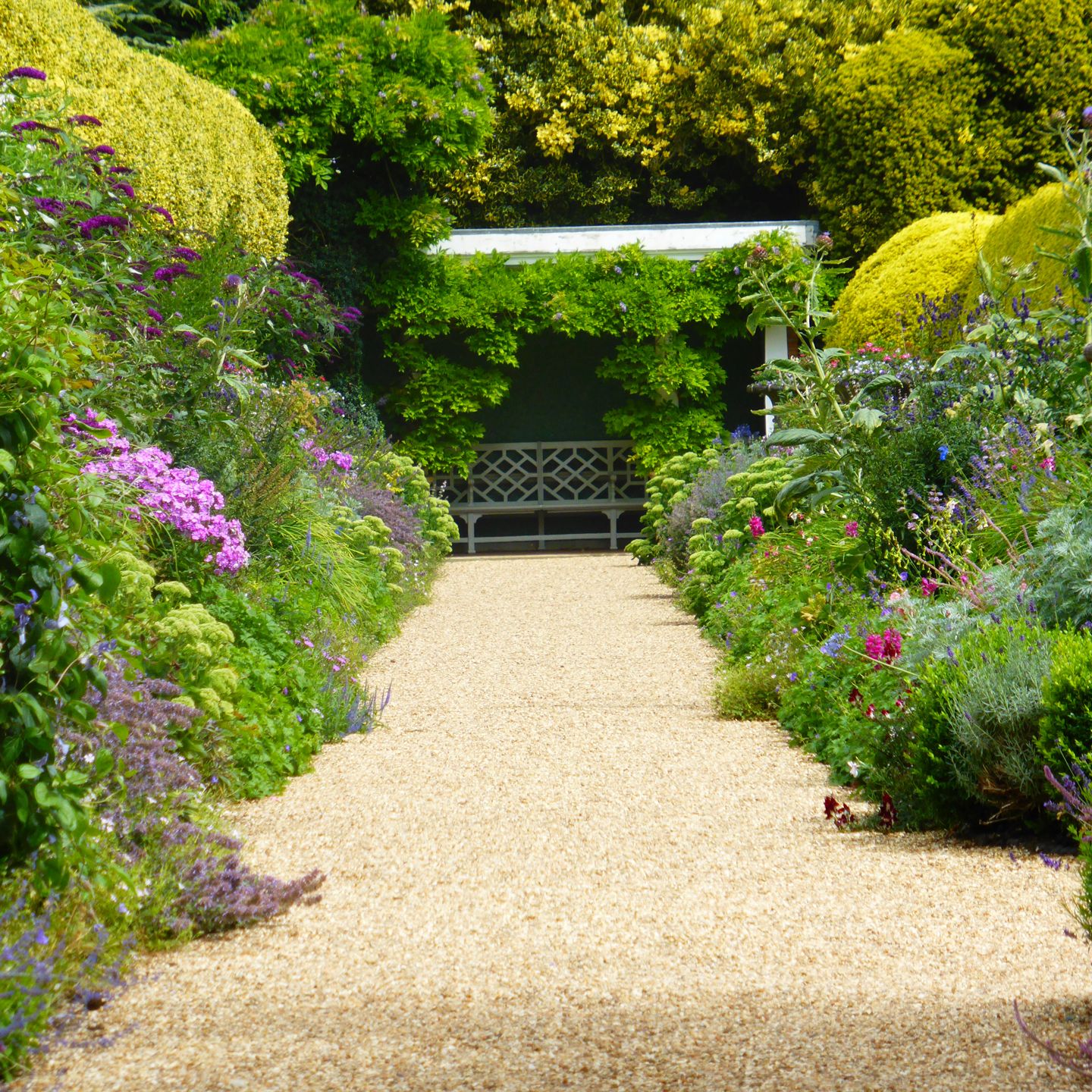 Pathway through a colorful garden with a bench under a leafy pergola